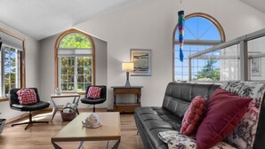 A spacious living room with a black leather sofa, two black chairs with pink cushions, a wooden coffee table, and a large window with a view of greenery.