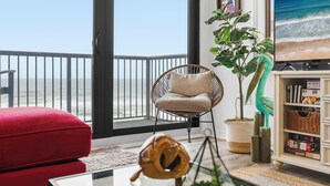 Uma varanda com vista para o oceano, uma cadeira de vime, um sofá vermelho, uma mesa de madeira e uma estante com livros e objetos decorativos.