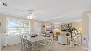 A spacious living area with a dining table, chairs, a ceiling fan, and a large window with blinds.
