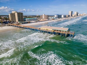 Ein Steg, der sich ins Meer erstreckt, mit einem Gebäude darauf, umgeben von einem Strand und Stadtgebäuden.