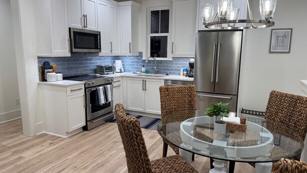 Una cocina moderna con armarios blancos, electrodomésticos de acero inoxidable y una mesa de comedor de vidrio rodeada de sillas de mimbre.