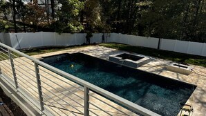 Piscina rectangular con solárium de piedra, rodeada por una cerca blanca, y un pequeño espacio de terraza con una fuente de agua.