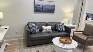 Una sala de estar con un sofá de cuero oscuro, dos almohadones decorativos, una mesa de centro blanca con una cesta tejida y un cuadro de un paisaje marino en la pared.