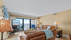 Ein Wohnzimmer mit braunem Ledersofa, einem Couchtisch, einem Fernseher und einem Balkon mit Blick aufs Meer.