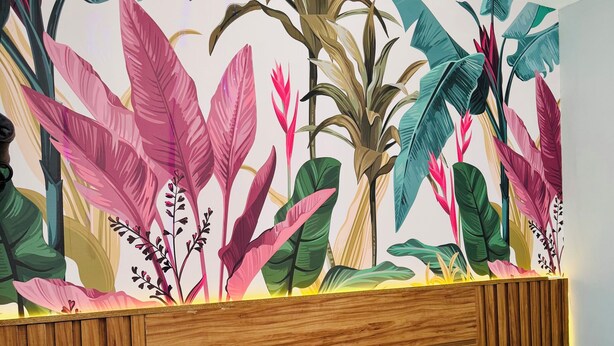 A bedroom with a bed, a wooden headboard, a wall mural of tropical leaves, a small stool, and a potted plant. - Prado Guest House (Florianópolis)