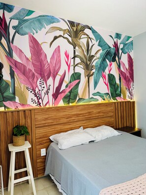A bedroom with a bed, a wooden headboard, a wall mural of tropical leaves, a small stool, and a potted plant. - Prado Guest House (Florianópolis)