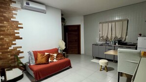 A modern living room with a sofa, a small table, and a kitchen area with cabinets and a window. - Prado Guest House (Florianópolis)