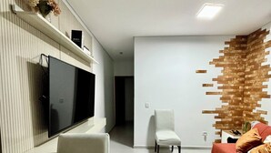 A modern living room with a flat-screen TV mounted on the wall, a wooden coffee table, a white sofa with cushions, and a decorative wall panel. - Prado Guest House (Florianópolis)