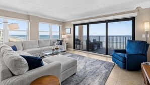 Un salon spacieux avec un grand canapé, un fauteuil bleu et une table basse en verre. Il y a un balcon avec vue sur l’océan.