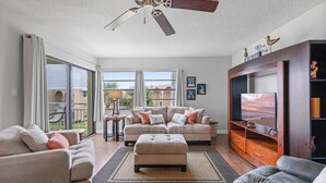 A spacious living room with a large window, a ceiling fan, a TV, and a wooden entertainment center. - Seaward B103 (New Smyrna Beach)