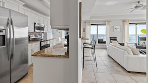 A modern kitchen with stainless steel appliances, a granite countertop, and a dining area with a white sofa and bar stools. - The Wave 302 (New Smyrna Beach)
