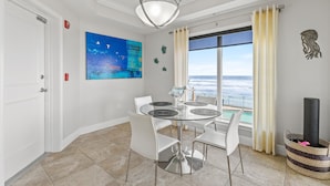 A dining area with a glass table, white chairs, a modern chandelier, a painting, and a view of the ocean. - The Wave 302 (New Smyrna Beach)