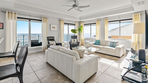 A spacious living room with a white sofa, a glass coffee table, and a ceiling fan. Large windows offer a view of the sea. - The Wave 302 (New Smyrna Beach)