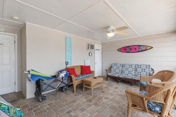 Una habitación con mobiliario de mimbre, ventilador de techo, tabla de surf colgada en la pared y un cochecito.