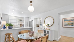Una cocina moderna con armarios blancos, una isla central y un comedor con mesa de vidrio y sillas acolchadas.