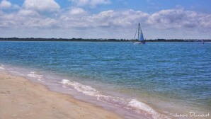 A beach with clear blue water, a sailboat in the distance, and a sandy shoreline.