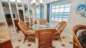 A dining area with wicker chairs and a glass table, large windows with a view of the ocean, and a chandelier.