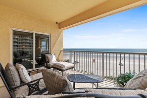 Un balcón con mobiliario exterior, una puerta corrediza de vidrio que da a un patio y vista a la playa.