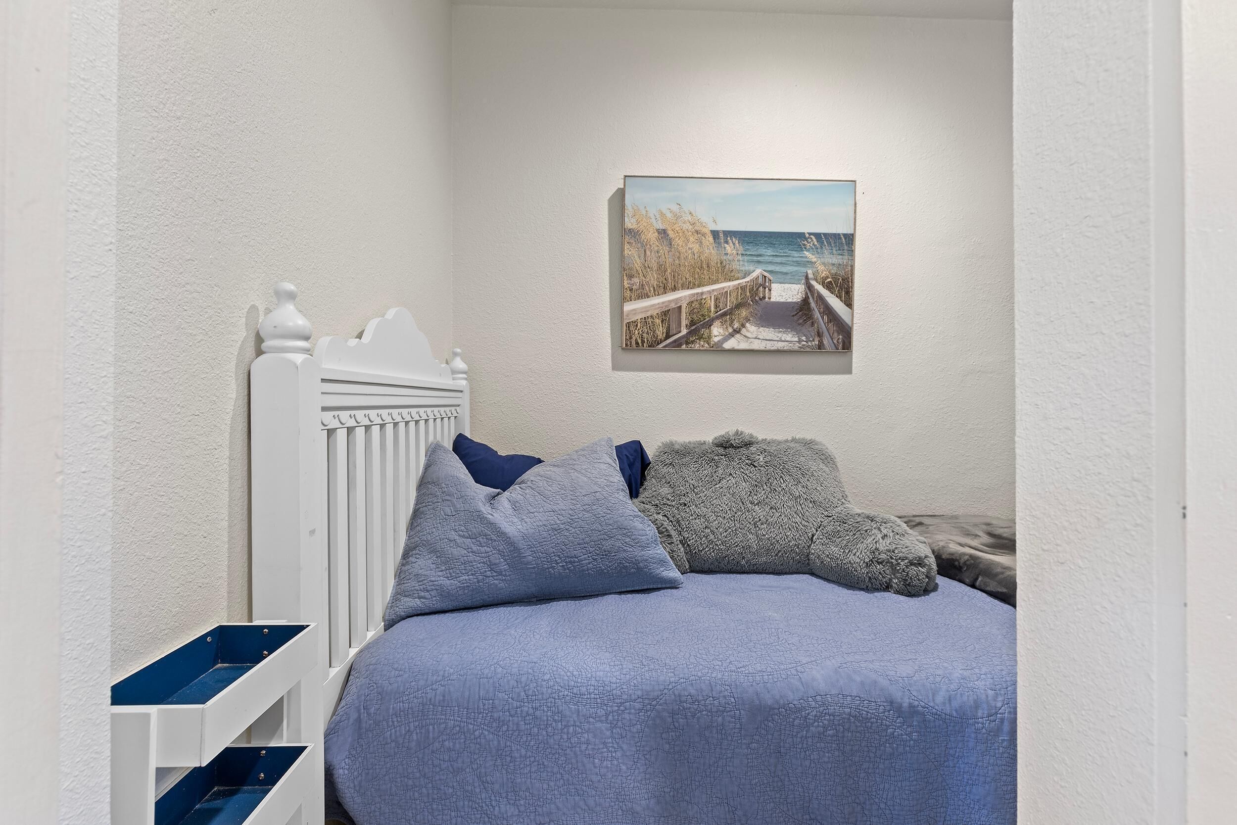 A bedroom with a white headboard, a blue bedspread, and a painting of a beach scene on the wall.