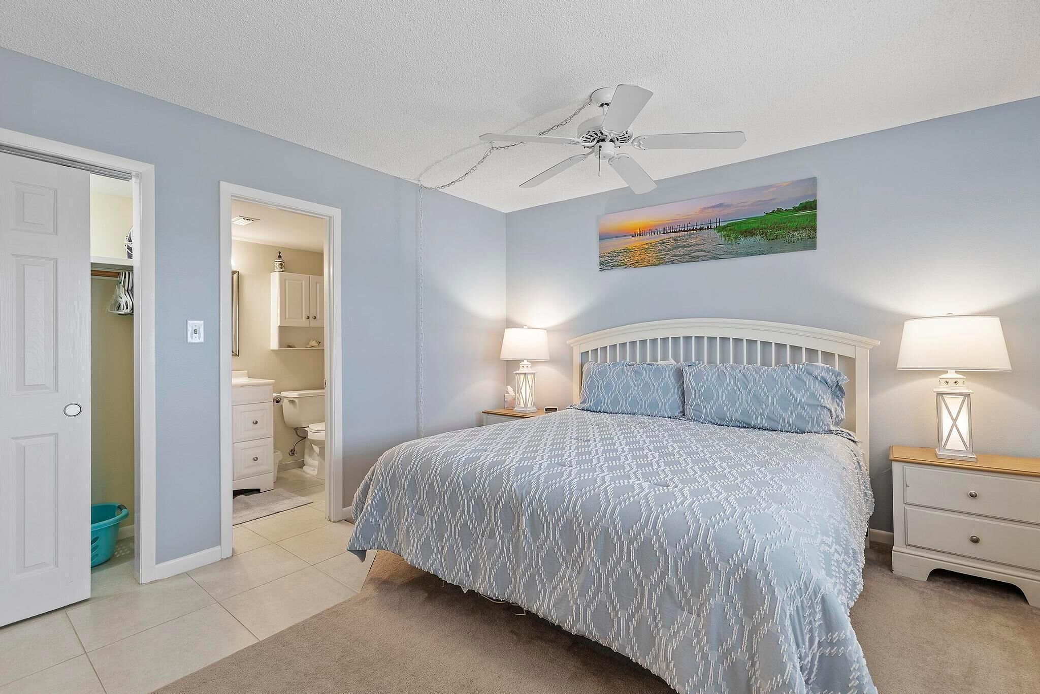 Une chambre à coucher avec un lit, des tables de chevet, un ventilateur de plafond, un tableau, et une salle de bain visible à travers une porte ouverte.
