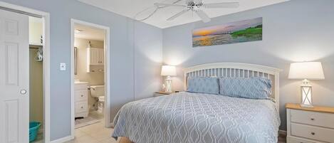 A bedroom with a bed, nightstands, a ceiling fan, a painting, and a bathroom visible through an open door.