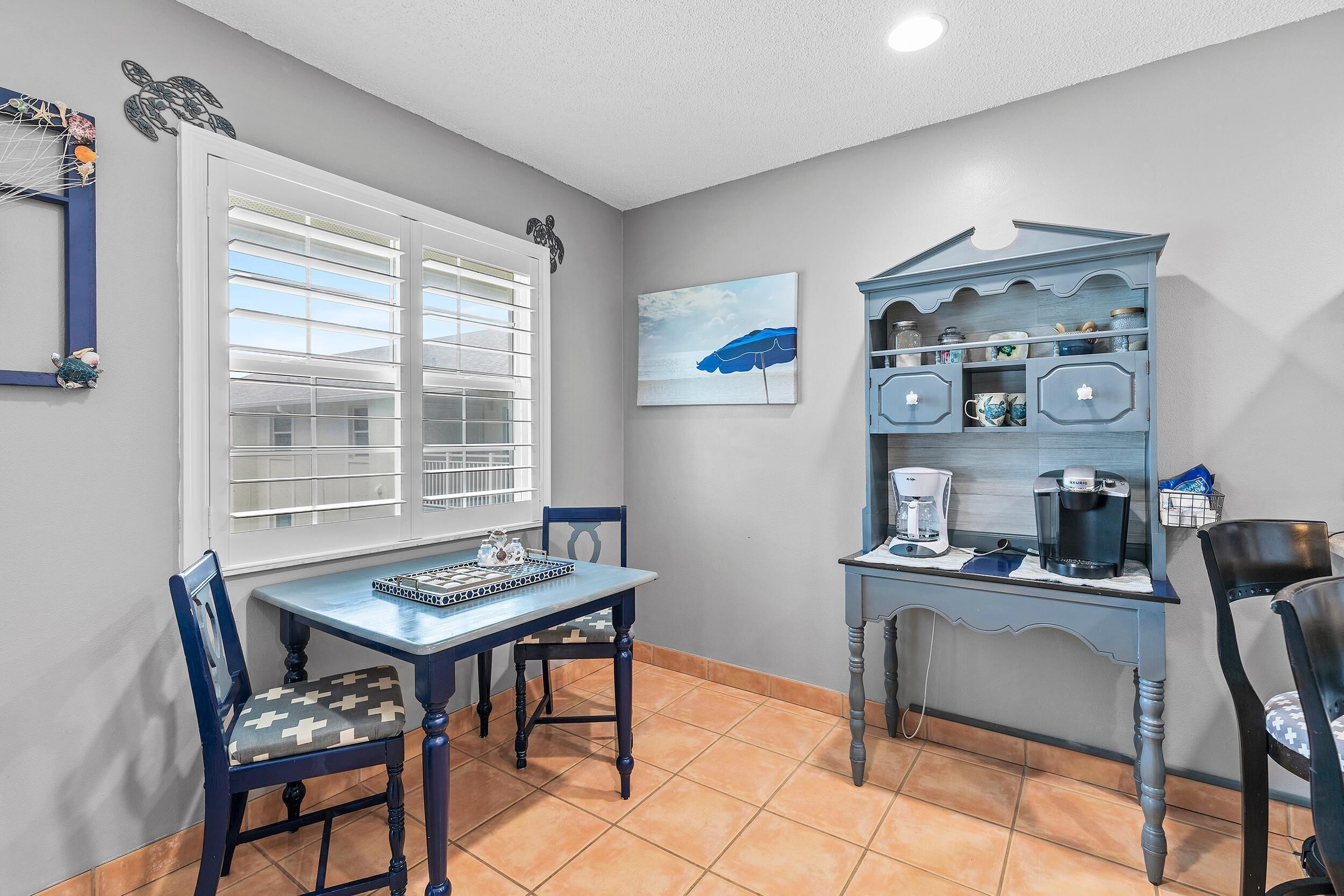 A dining area with a table, chairs, a coffee station, and a window with blinds.