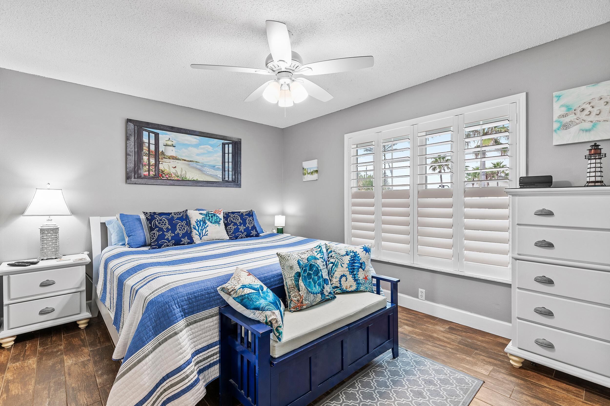 A bedroom with a bed, nightstands, a dresser, a ceiling fan, and a window with shutters.