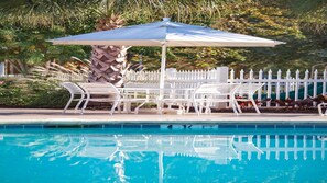 Ein Poolbereich mit weißen Liegestühlen und einem Sonnenschirm, umgeben von einem weißen Zaun und Palmen.