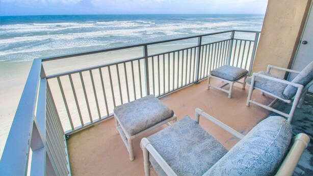 A balcony with two chairs and a small table, overlooking a beach with waves.