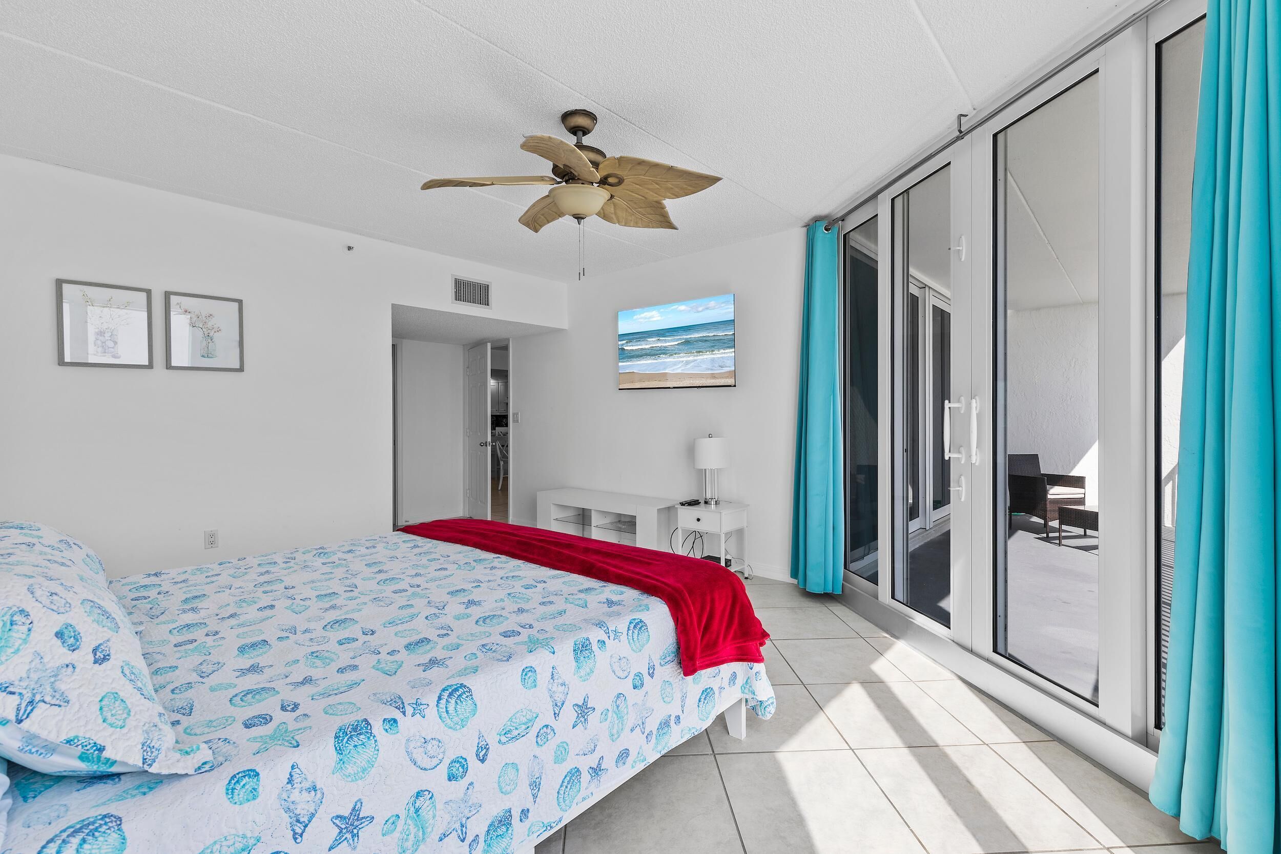 A bedroom with a bed, a ceiling fan, a TV, and a sliding glass door to a balcony.