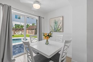 Een eetgedeelte met een witte tafel, vier stoelen, een vaas met bloemen en een groot raam met uitzicht op een zwembad en tuinmeubilair.