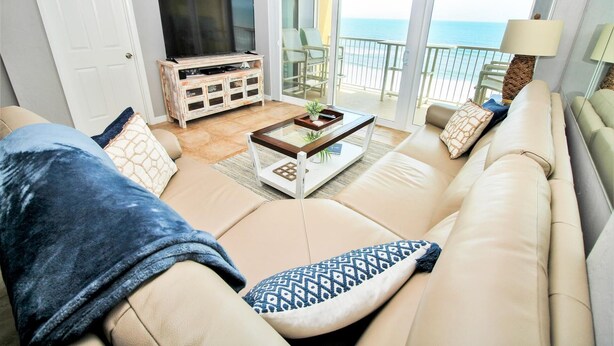 Ein Wohnzimmer mit beiger Couch, Couchtisch und Fernseher auf einem Holzboden. Ein Balkon bietet Blick aufs Meer.