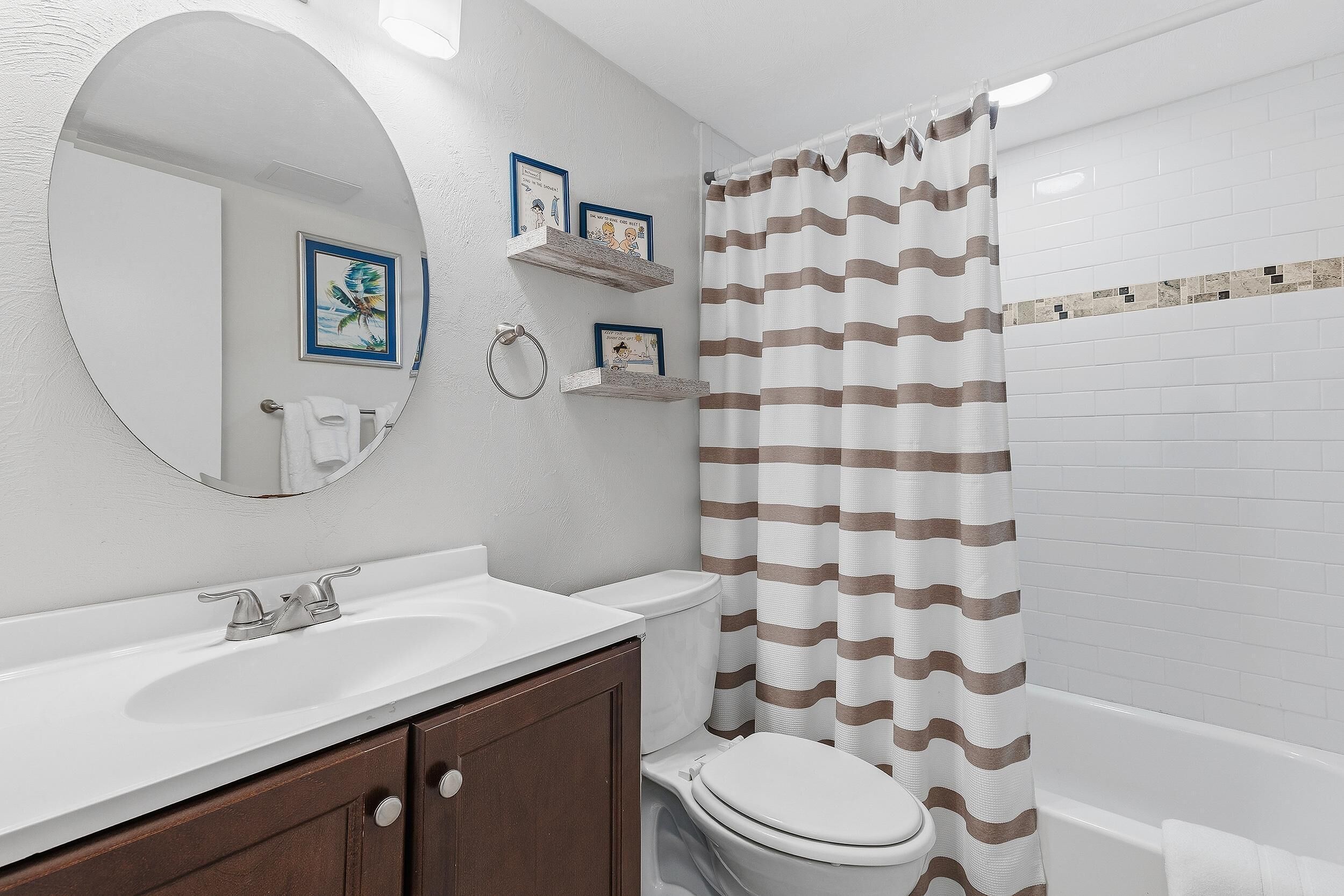 A bathroom with a white sink, a toilet, a shower curtain with a striped pattern, and a mirror with a circular frame.