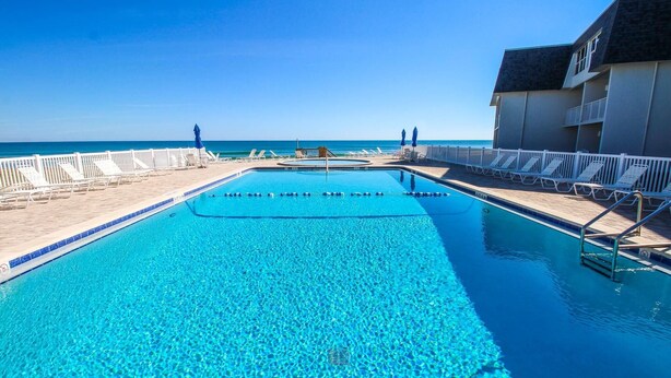 A clear blue pool with a view of the ocean, surrounded by white lounge chairs and umbrellas. - Surfside 107 (New Smyrna Beach)