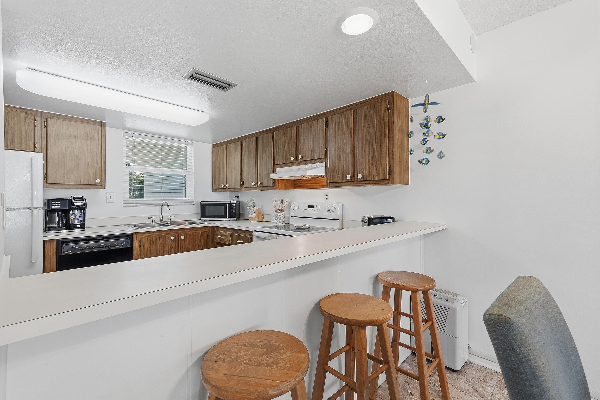 Cocina moderna con armarios de madera, encimera blanca, horno empotrado, microondas y cafetera. Hay dos taburetes de madera y una ventana con persianas.
