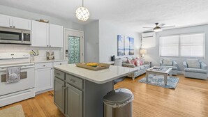A modern kitchen with white cabinets, a microwave, and a stove. There's a dining area with a table and chairs, and a living room with a couch and a ceiling fan.