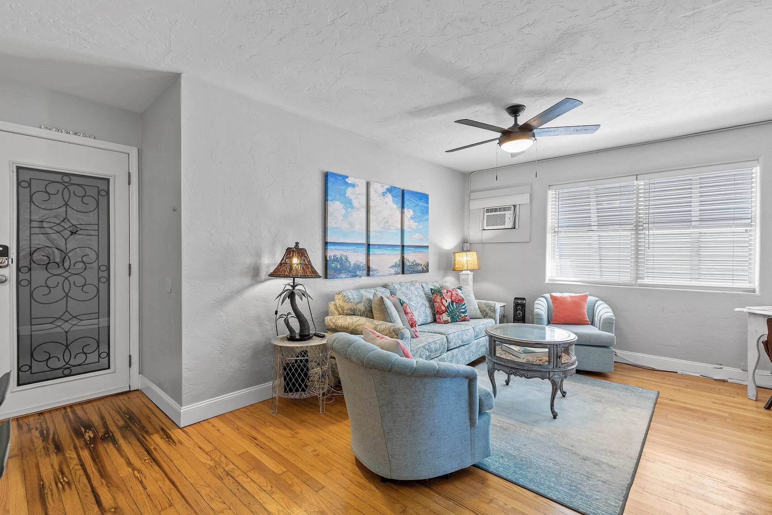 A living room with a blue armchair, a glass coffee table, a ceiling fan, and a large window with blinds.