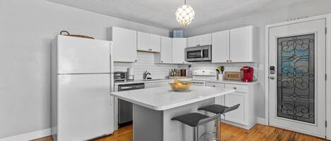 A modern kitchen with white cabinets, a central island with stools, a microwave, and a hanging light fixture.