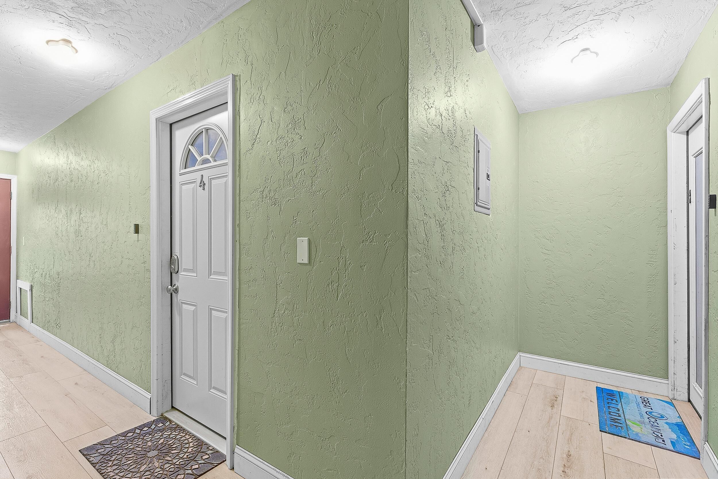 Un pasillo con pared verde con textura, una puerta blanca con panel de vidrio y un tapete azul en el suelo.