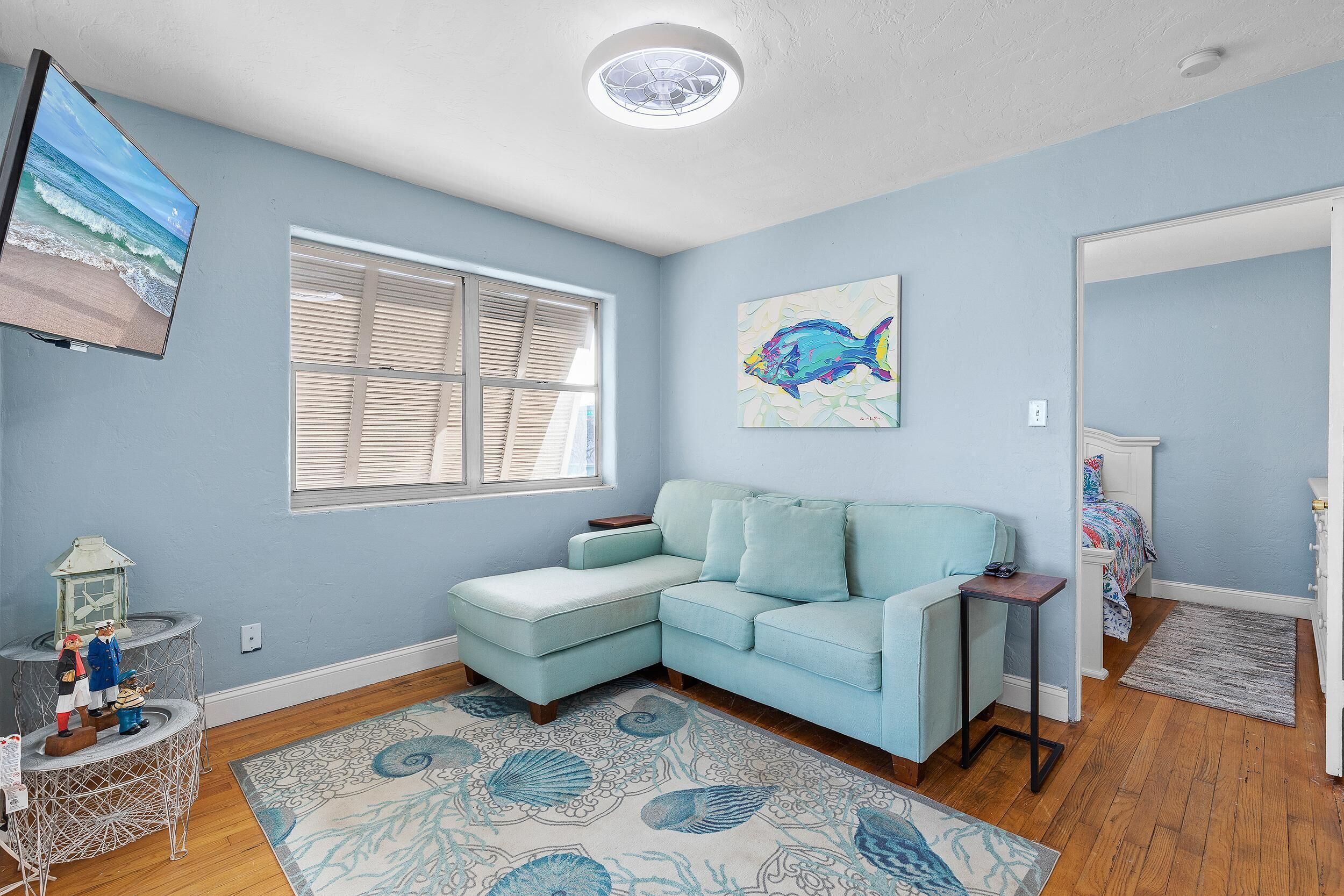 A room with a light blue wall, a large window with blinds, a teal sofa, a small side table, a decorative rug, and a wall-mounted TV.