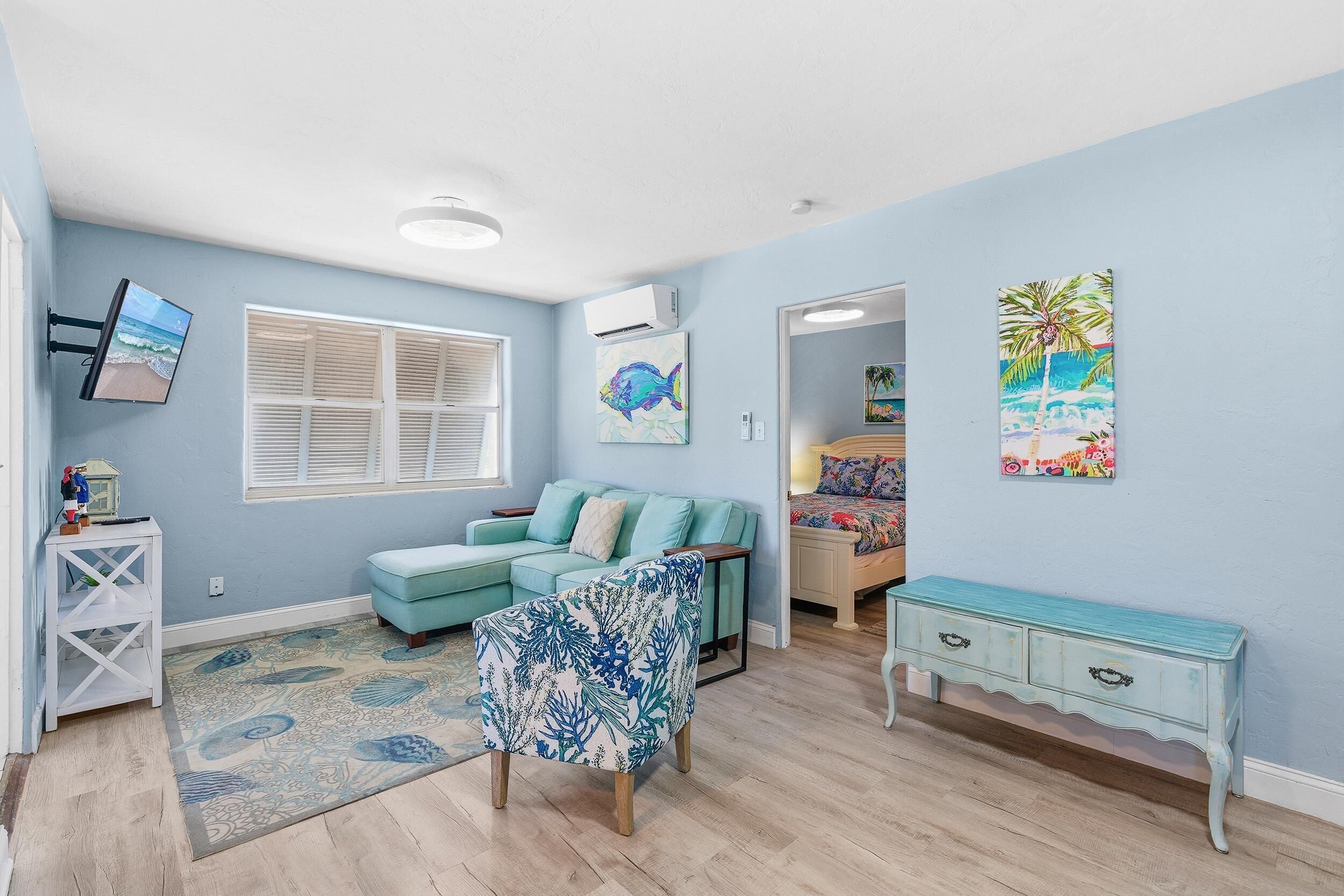 A room with a light blue wall, a teal sofa, a patterned armchair, a white console table, and a TV mounted on the wall.