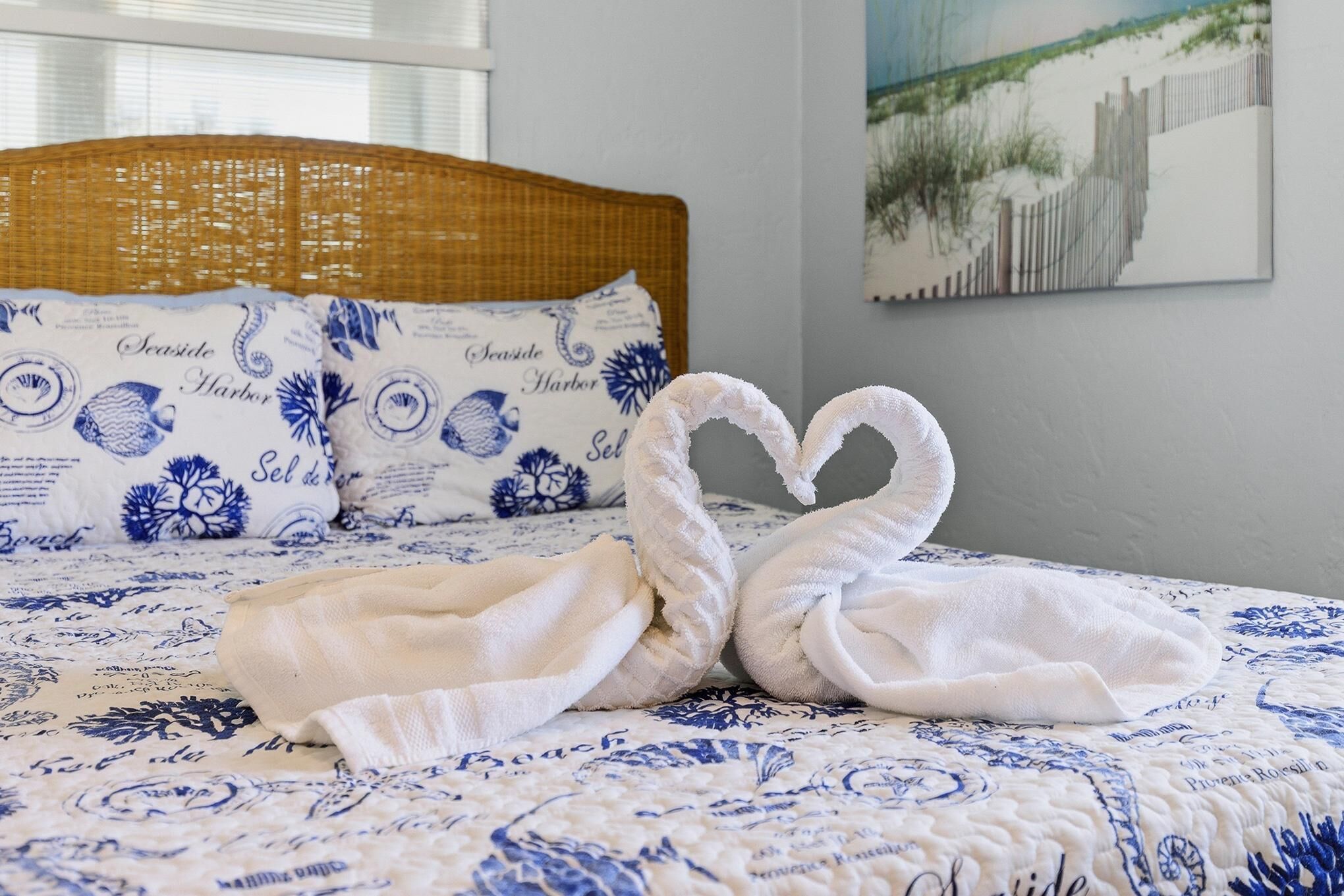Een bed met een blauw bloemenbedekking, twee handdoeken gevouwen in de vorm van een hart, en een rieten hoofdbord.