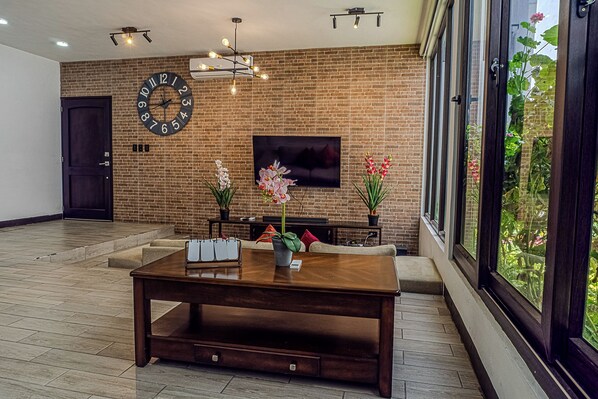 A modern living room with a brick wall, a large clock, a flat-screen TV, a wooden dining table, and a view of greenery through large windows. - Hermosa casa, con Parqueo propio, Ideal para pasar momentos en familia o pareja. (Jocotenango)