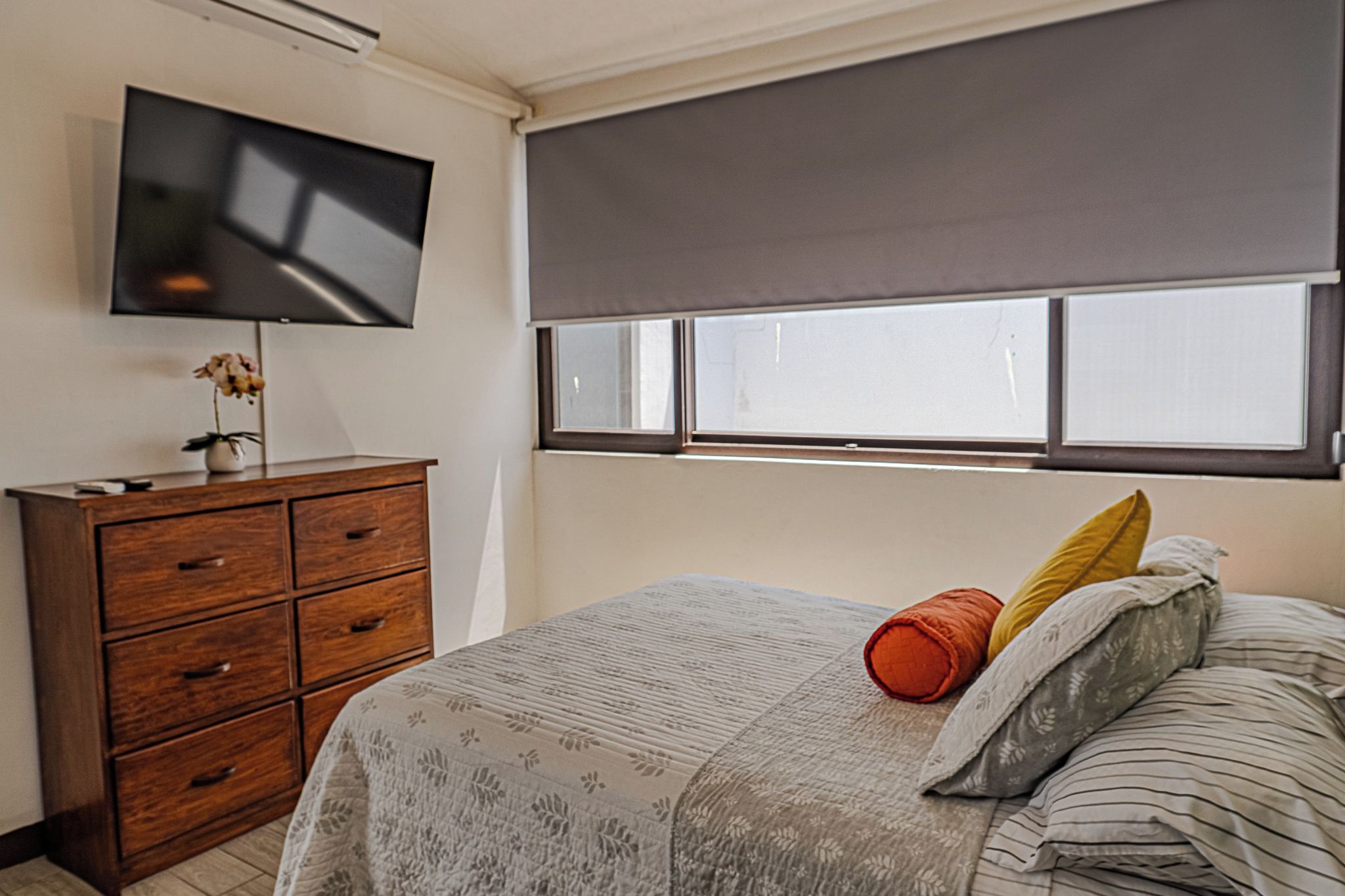A bedroom with a bed, a wooden dresser, a television on the wall, and a window with blinds.
