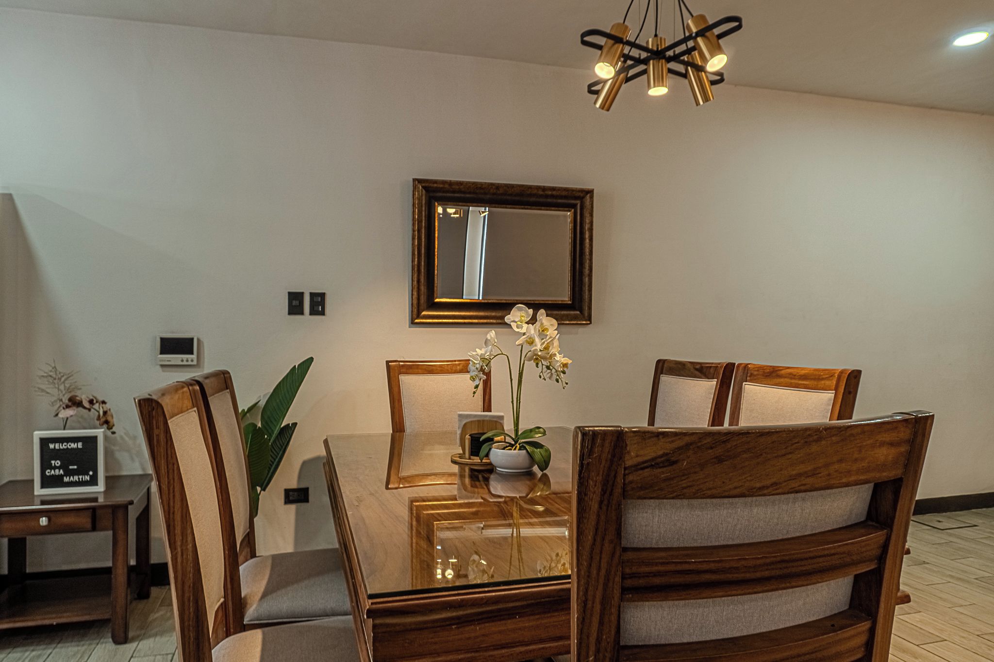 A dining area with a wooden table, chairs, a chandelier, a mirror, and a welcome sign.