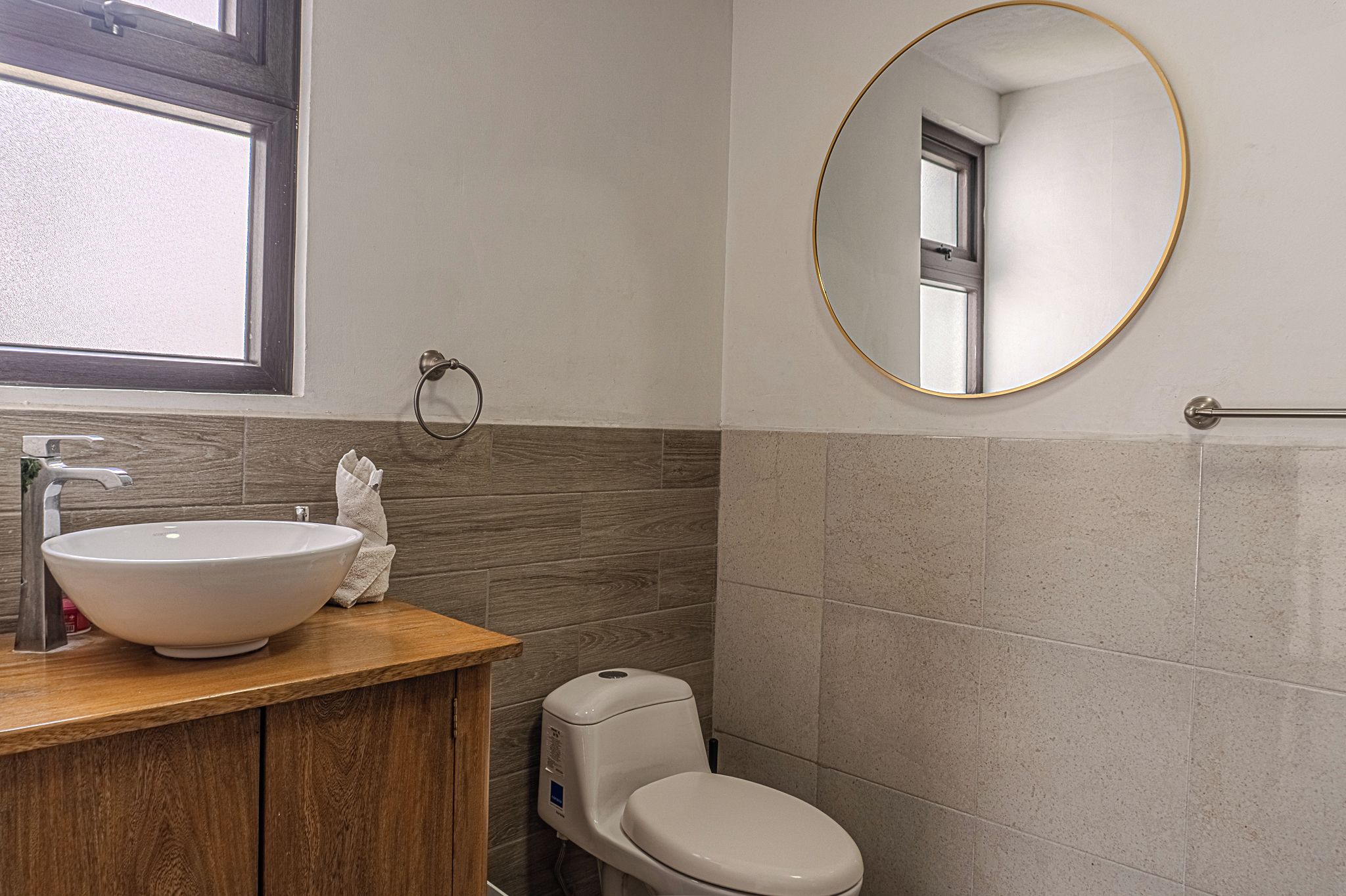 A bathroom with a wooden vanity, a white sink, a toilet, and a round mirror with a gold frame.