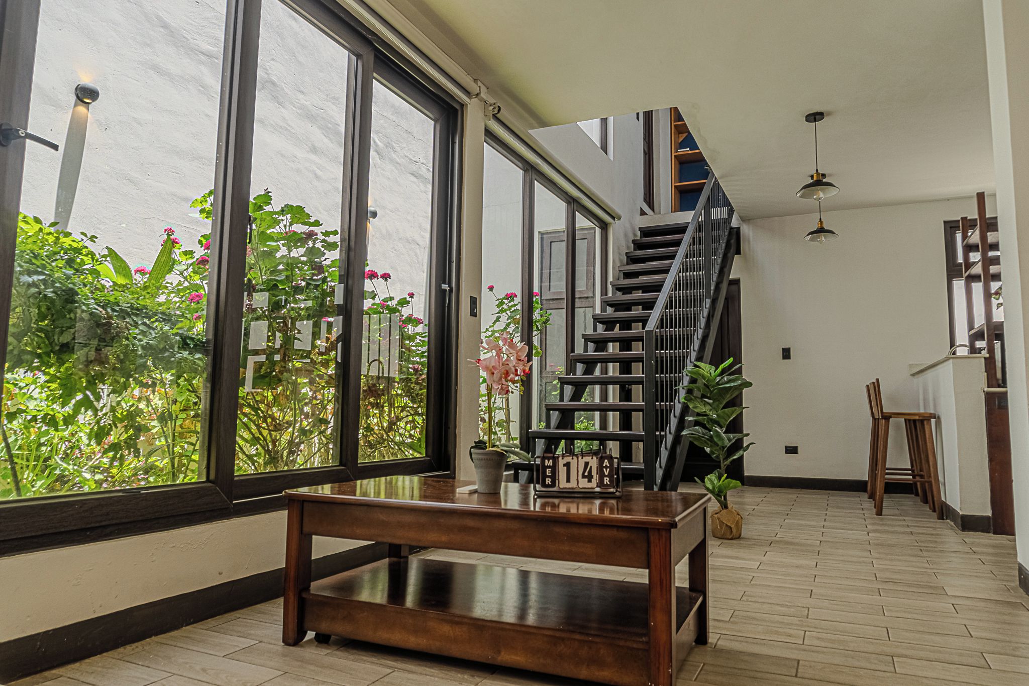 A spacious room with a wooden table, a staircase, and large windows overlooking a garden.