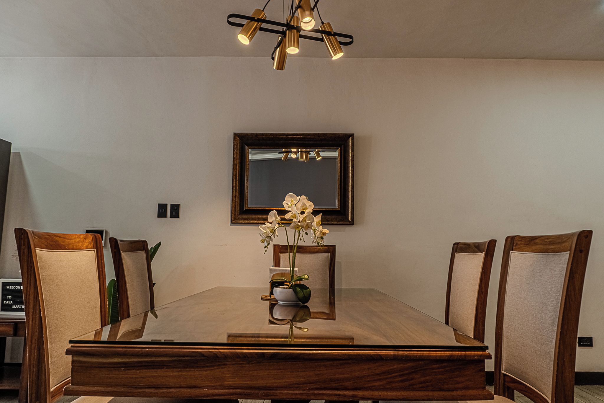 Une salle à manger avec une table en bois, des chaises, un vase de fleurs, un miroir au mur et un lustre.