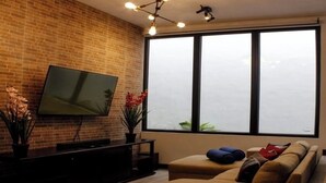 A living room with a brick wall, a flat-screen TV, a sofa, and a large window with a view. - Hermosa casa, con Parqueo propio, Ideal para pasar momentos en familia o pareja. (Jocotenango)