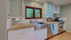 Une cuisine moderne avec des meubles blancs, un évier en acier inoxydable, un lave-vaisselle, un four et une fenêtre donnant sur un paysage verdoyant.